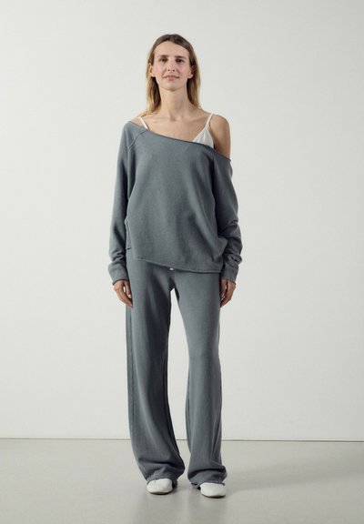 Femme debout portant un pantalon large en maille grise, un pull gris oversize tombant sur une épaule laissant apparaître un caraco blanc, avec des chaussures blanches sans lacets.