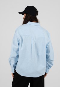 Femme portant une chemise à manches longues rayée bleu clair et blanc et une casquette de baseball noire, dos à la caméra.