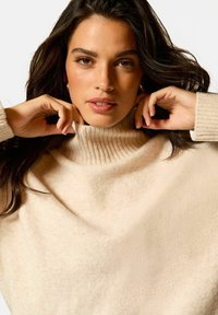 Sweter z golfem w kolorze kremowym, z ribbowanym kołnierzem, miękką teksturą i luźnym krojem. Modelka poprawia kołnierz obiema rękami.