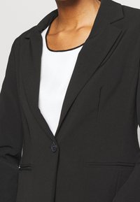 Blazer nero su misura con colletto a revers e chiusura a un bottone. Tessuto liscio, due tasche laterali e una camicia bianca sotto.