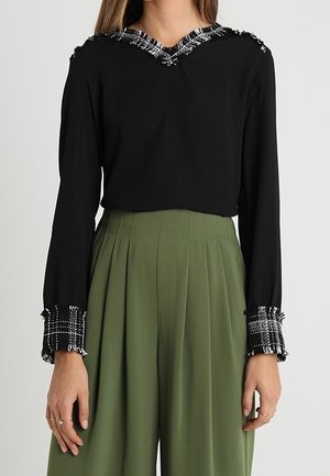 Czarna bluzka z długim rękawem z czarno-białą, kratkowaną lamówką z frędzlami na dekolcie w kształcie litery V i mankietach, połączona z oliwkowozielonymi plisowanymi spodniami z wysokim stanem.