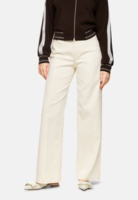 Weite, cremefarbene Hosen aus Baumwolle. Sie haben eine hohe Taille und einen geraden Schnitt, kombiniert mit off-weißen Schuhen mit Schleifenakzenten.