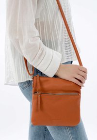 Oranžová kožená crossbody taška s texturovaným povrchem, která má zipovou kapsu a dlouhý nastavitelný popruh. Kombinována s bílým topem.