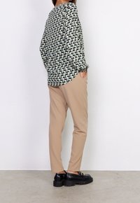 Camicia a maniche lunghe con un motivo geometrico in verde, nero e crema; abbinata a pantaloni beige e scarpe con plateau nere.