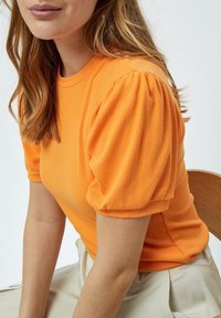 Oranžs ribots adīts tops ar īsām pūkainām piedurknēm, apaļu kakla izgriezumu un pieguļošu siluetu. Audumam ir gluda tekstūra.