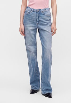 Vrouw die lichtblauwe rechte jeans draagt met een roze top en zwarte puntige schoenen, staand tegen een egale achtergrond.
