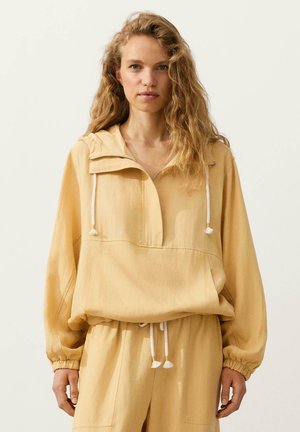 Młoda kobieta z długimi kręconymi blond włosami, ubrana w luźną, beżową bluzę z kapturem i dopasowane spodnie na jednolitym jasnym tle.