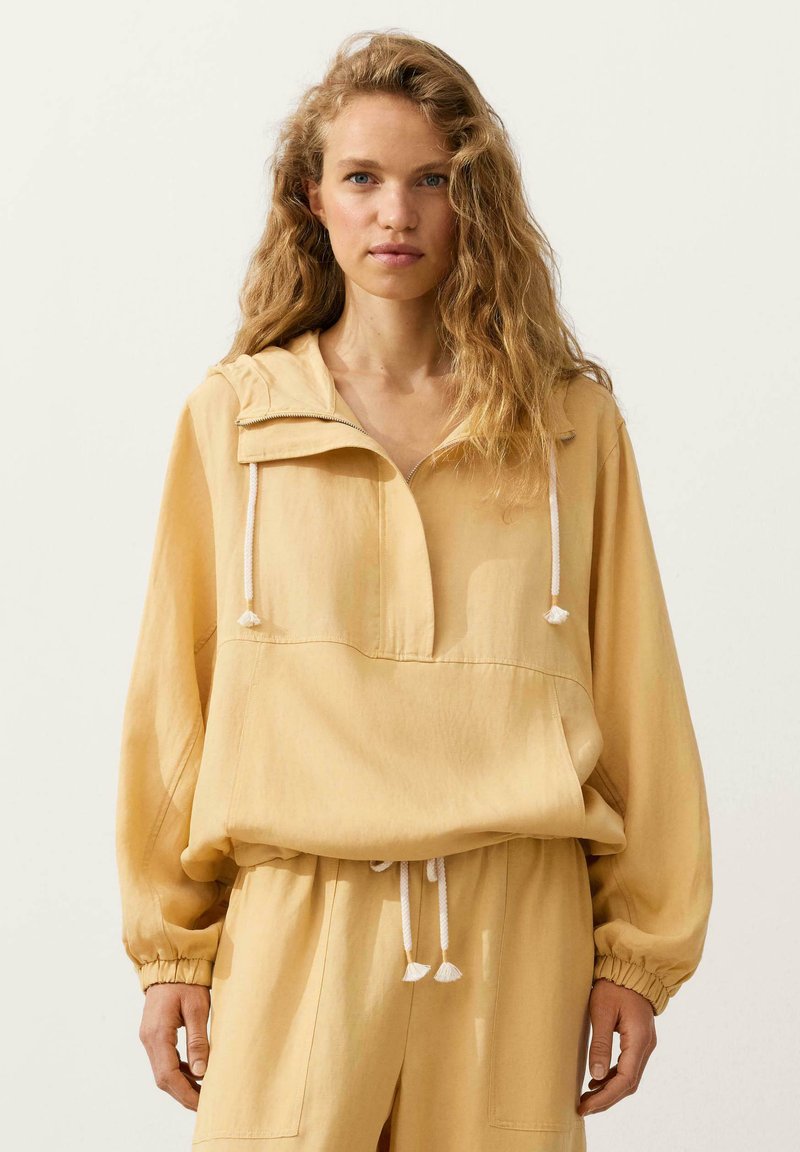Jeune femme aux longs cheveux blonds bouclés, portant un sweat à capuche beige ample et un pantalon assorti sur un fond clair uni.