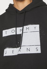 Černá mikina s kapucí a s tkaničkami, na které jsou bílé obdélníkové bloky a text "TOMMY JEANS" vytištěný přes hrudník.