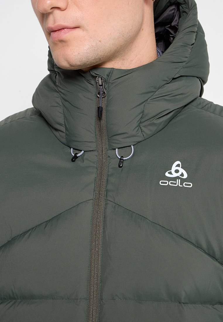 Giacca verde imbottita con colletto alto e cappuccio. Presenta chiusura con zip, logo sul petto e texture trapuntata.