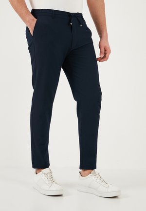 Muž nosi tmavomodré slim-fit nohavice so sťahovacou šnúrkou v páse a biele tenisky, stojí s jednou rukou v vrecku.