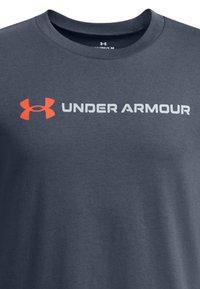 Camiseta de manga corta gris oscuro hecha de algodón. Presenta un prominente logo naranja de Under Armour y texto gris. Diseño de cuello redondo.