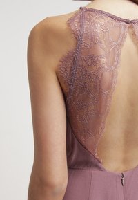 Vue arrière d'une robe avec un décolleté en V profond, en tissu mauve doux, et un détail en dentelle intricate le long des épaules et de l'encolure.