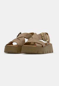 Beige cross-strap sandals with a chunky, textured platform sole. Gold buckle detail enhances design. Soft, suede-like material.