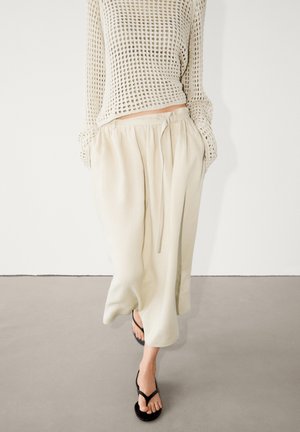 Femme portant un pantalon beige ample à taille nouée, un pull en maille crème à manches ouvertes, et des sandales noires à enfiler, marchant sur un sol gris clair.