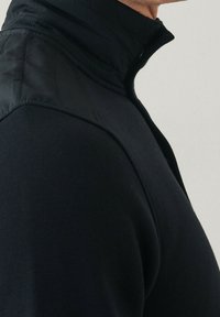 Uomo che indossa una giacca nera con zip e spalle imbottite, mostrato di lato e ritagliato sopra il mento su uno sfondo chiaro.