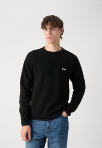 Czarny sweter dzianinowy z okrągłym dekoltem, ściągaczami na rękawach i małym białym logo 'BOSS' na piersi; zestawiony z niebieskimi dżinsami.