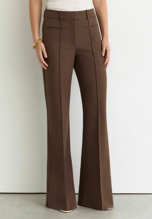 Frau trägt hoch taillierte braune, weit geschnittene Hose mit Nahtdetails, kombiniert mit einem weißen Top und einem massiven goldenen Armband am linken Handgelenk.