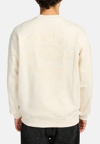 Felpa color crema con logo e testo in rilievo sul retro, scollatura a girocollo e polsini e orlo a coste. Materiale in cotone morbido.