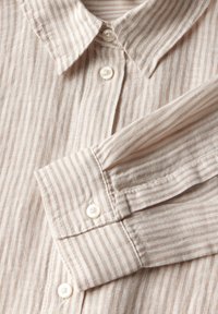 Camisa de lino color beige claro con finas rayas blancas, que cuenta con un cuello clásico, botones de perla y mangas largas con puños abotonados.