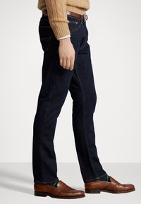 Vaqueros de mezclilla azul oscuro con un corte recto, costuras detalladas, combinados con mocasines de cuero marrón y un suéter beige de punto grueso con trenzas.