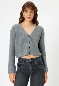 Grijze cropped cardigan met een V-hals, geribbelde textuur, lange mouwen en vijf zwarte knopen aan de voorkant, gecombineerd met zwarte jeans.