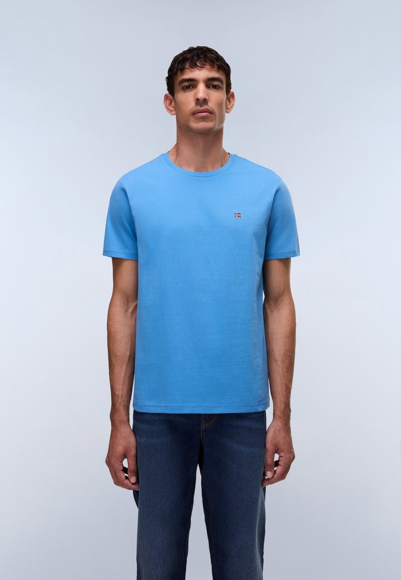 Mann trägt ein einfarbiges blaues T-Shirt mit kurzen Ärmeln und kleinem Flaggenemblem auf der Brust sowie dunkelblaue Jeans, steht vor einem hellgrauen Hintergrund.