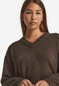 Brauner Oversized-Pullover mit V-Ausschnitt aus strukturiertem Strickstoff; mit gerippten Bündchen und Halsauschnitt; das Modell hält die Hände in der Taille.