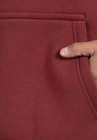 Tela burdeos con una textura suave, que presenta un bolsillo lateral y detalles de costura visibles. Una mano está posicionada cerca de la abertura del bolsillo.