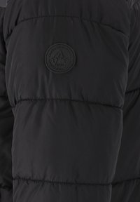 Sort pufferjakkeærme med en quiltet design, glat tekstur og en cirkulær sort lap med prægede tekst- og logodetaljer.