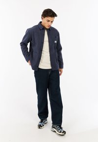 Jonge man in een marineblauwe jas over een wit shirt, donkere jeans en zwart, grijs en witte sneakers, staande tegen een witte achtergrond.