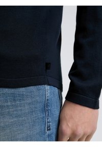 Maglione a maglia navy con maniche lunghe, polsini a coste e una piccola etichetta con logo sul lato, abbinato a jeans in denim blu chiaro.