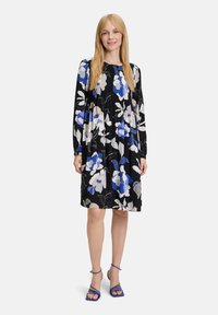 Blumenkleid aus schwarzem Stoff mit weißen, blauen und grauen Blumen. Lange Ärmel und A-Linien-Schnitt, kombiniert mit blauen Riemchensandalen.