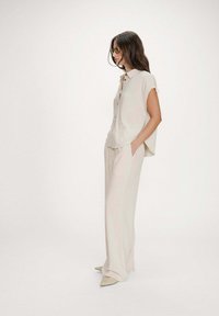 Mujer vistiendo una blusa sin mangas color beige con botones, pantalones de pierna ancha, zapatos puntiagudos y gafas de sol, de pie en un fondo de estudio minimalista.