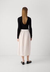 Donna con capelli lunghi e lisci, che indossa una maglietta nera a maniche lunghe, una gonna midi bianca e delle ballerine nere, in piedi di fronte a un muro bianco semplice.