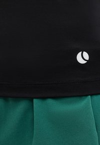 Μαύρο υφασμάτινο με υφή και μικρό λευκό κυκλικό λογότυπο πάνω από λείο πράσινο ύφασμα, που δείχνει λεπτομέρειες ενδύματος και χρωματική αντίθεση.