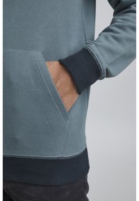 Sudadera de un azul claro con puños y dobladillo de un azul oscuro acanalado, que cuenta con un bolsillo canguro y una textura suave. Las costuras son visibles.