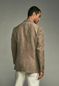 Blazer en daim marron clair avec une fente unique à l'arrière, revers crantés et boutons noirs sur les poignets, associé à un pantalon couleur crème.