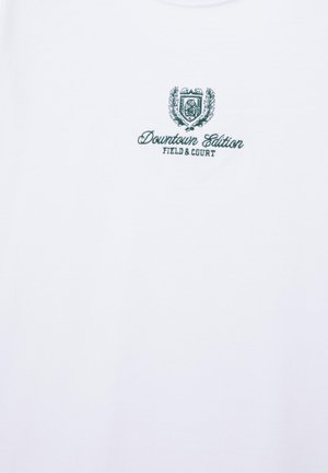 Wit overhemd met klein groen geborduurd schild boven de tekst "Downtown Edition Field & Court" in het midden van de borst.