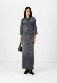 Denim maxikjole i mørkegrå, med en knappestolpe foran, to brystlommer, og belte i livet. Stylet med svarte ankelstøvler og en håndveske.