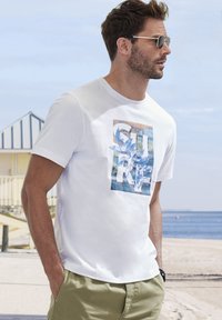 Weißes T-Shirt mit kurzen Ärmeln, das ein blau-grünes Grafikdesign mit dem Text "SURF" zeigt. Das Model trägt eine Sonnenbrille und olivgrüne Shorts.