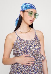 Vestido sem mangas com padrão floral em bege e roxo, com um laço na cintura. Acessorizado com um lenço de cabeça azul com padrão e óculos de sol verdes.