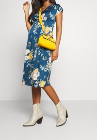 Nainen, jolla on yllään sininen kukkamekko, vaaleat nilkkasaappaat ja kirkas keltainen olkalaukku, poseeraa yksinkertaista taustaa vasten.