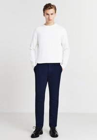 Jeune homme debout avec les mains dans les poches de son pantalon marine, portant un pull à manches longues blanc et des chaussures habillées noires sur un fond clair.