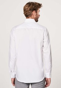 Chemise blanche à manches longues avec un col classique, texture lisse et poignets à boutons, présentant un ourlet arrondi en bas.