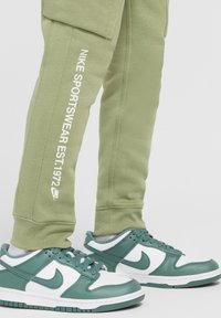 Pantaloni jogger verdi in cotone con testo bianco "NIKE SPORTSWEAR EST.1972" e dettagli verdi abbinati, indossati con sneakers Nike bianche e verdi.