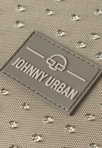 bézs szövet, textúrával, kis gyémántcsiszolt kövekkel és szürke gumi címkével, amelyen a "JOHNNY URBAN" felirat van emboszolva.