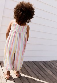 Jumpsuit pastellato a righe con corpetto arricciato e polsini alla caviglia arricciati; tessuto leggero. Indossato con sandali rosa chiaro, visto da dietro.