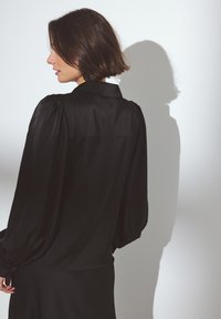 Blusa nera in satin con colletto, maniche a palloncino e polsini arricciati, dal taglio sagomato e dalla texture liscia.