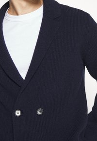 Námorně modrý pletený cardigan s šálovým límcem, zav�íráním na dvojité knoflíky a žebrovanou texturou. Přes bílou košili pod ním.